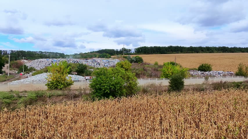 Golden cornfield stretches under a cloudy sky. Rubble piles rise behind green shrubs and trees. Faint horizon shows distant forest and rolling hills
