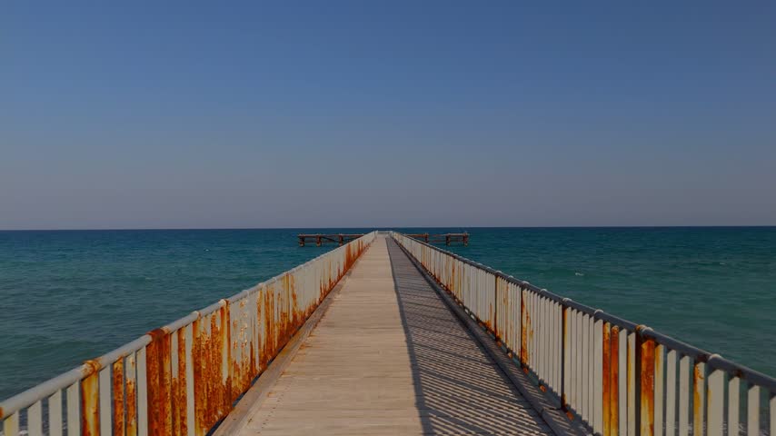 An aerial low-altitude tracking shot follows a wooden pier leading into the Mediterranean Sea at Mavralis Beach, ending with a pedestal move and tilt down over turquoise water.