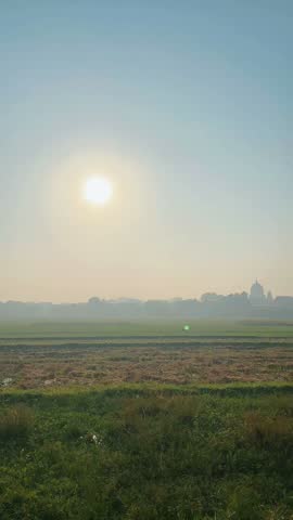 A tranquil dawn scene showcasing a hazy sun glowing over agricultural fields with a distant silhouette of a village and a mosque dome under a misty sky.