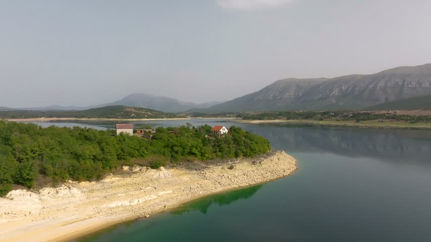 Small cluster of red-roof homes sits on a tree-covered peninsula of Peruća Lake with exposed rocky shore and hazy mountain backdrop.