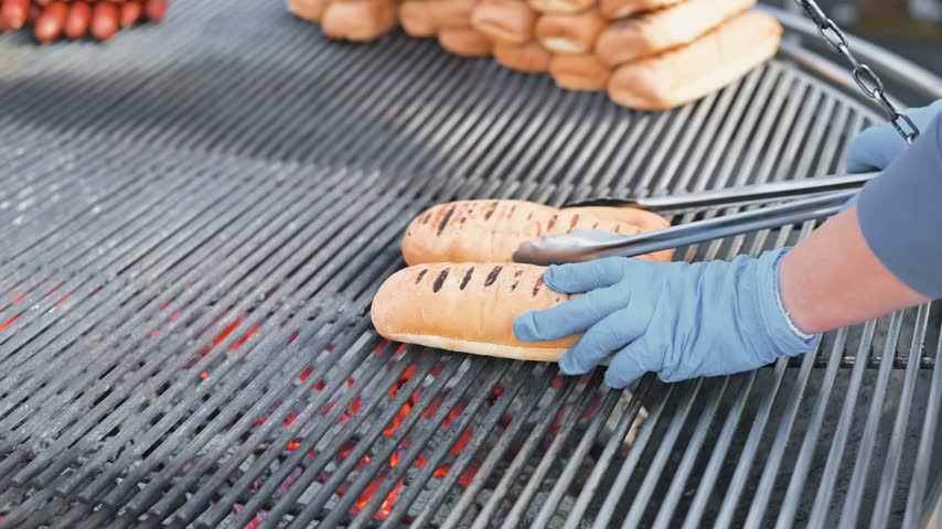 Hot dog buns are placed on a grill, showing grill marks as they cook. Smoke rises from the grill, indicating a barbecue setting where food is being prepared for serving.