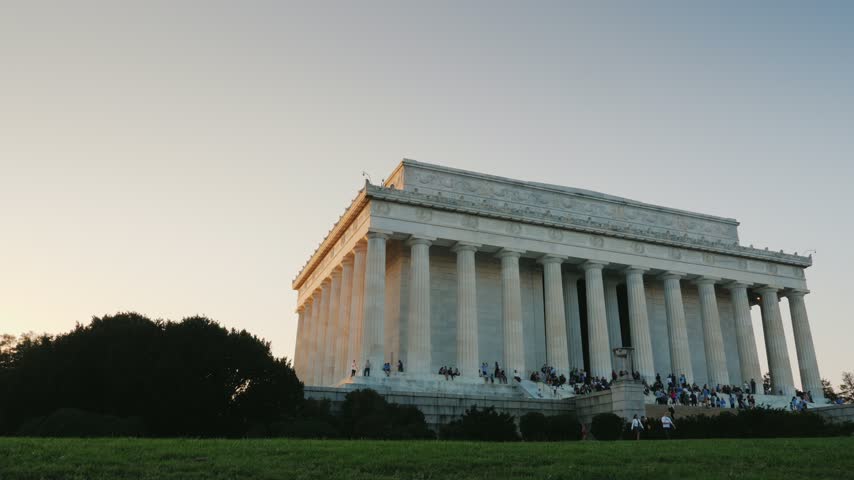 Stunning low angle shot of historic Lincoln Memorial in Washington DC during golden hour with tourists gathered on steps against clear sky cinematic footage capturing iconic landmark atmosphere.