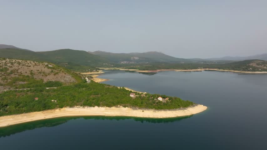 Sandy peninsula with a few forest-backed homes curves into a wide reservoir bay beneath low green hills, viewed from a moving drone.