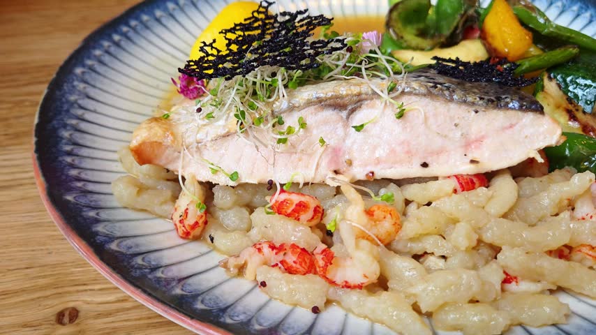 close view shows salmon with crispy seafood pasta and grilled vegetables, garnished with sprouts, flowers and seaweed on a patterned ceramic plate