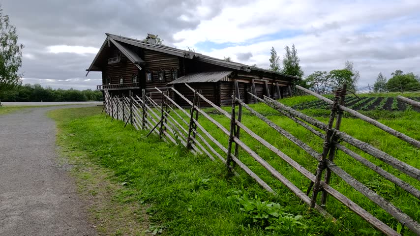 Old wooden house stands alone in a grassy field under a cloudy sky, with a gravel path and a weathered wooden fence in front of it. Overcast light creates a quiet, slightly melancholic rural scene.