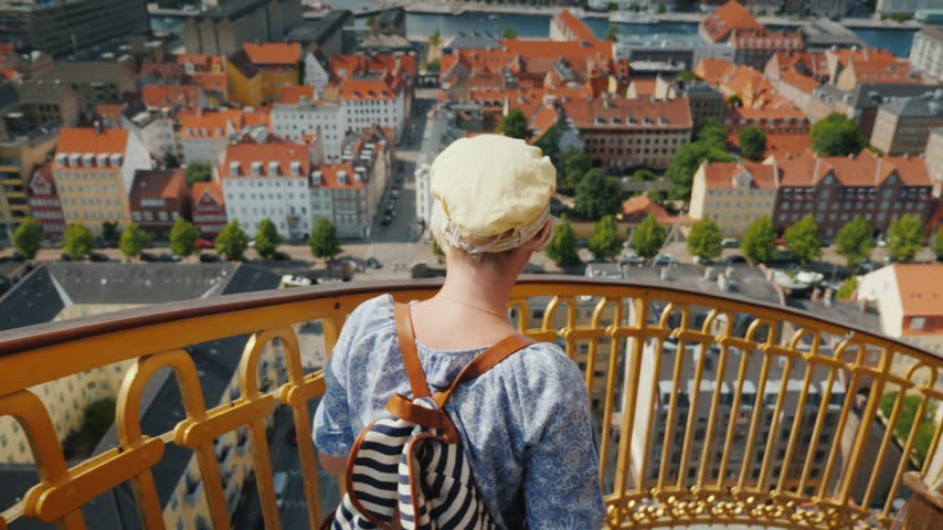 Young female traveler wearing cap and backpack standing on Church of Our Saviors golden balcony enjoying panoramic views over Copenhagen city historic center during sunny summer day.