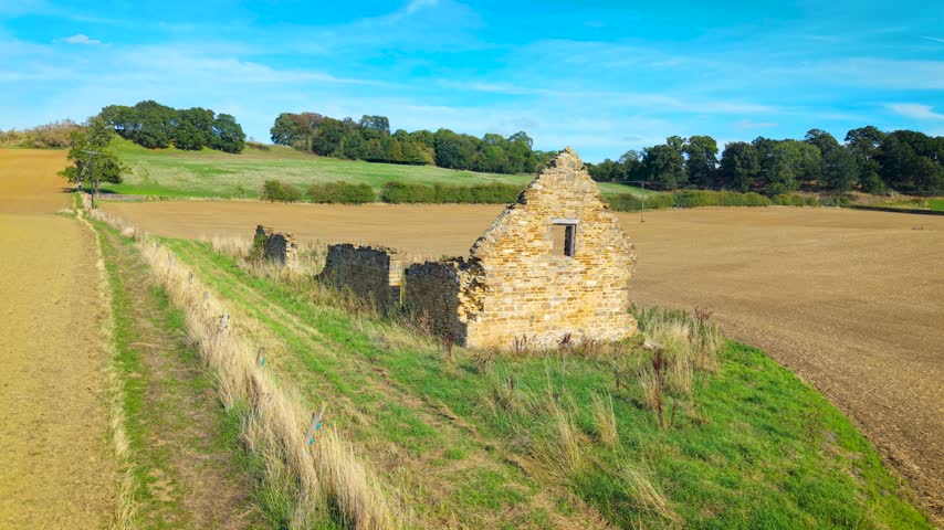 Ancient stone farmhouse ruins beside cultivated fields. Weathered gable wall with narrow window along grassy farm track. Rural agricultural landscape with masonry remains, hedgerows and rolling