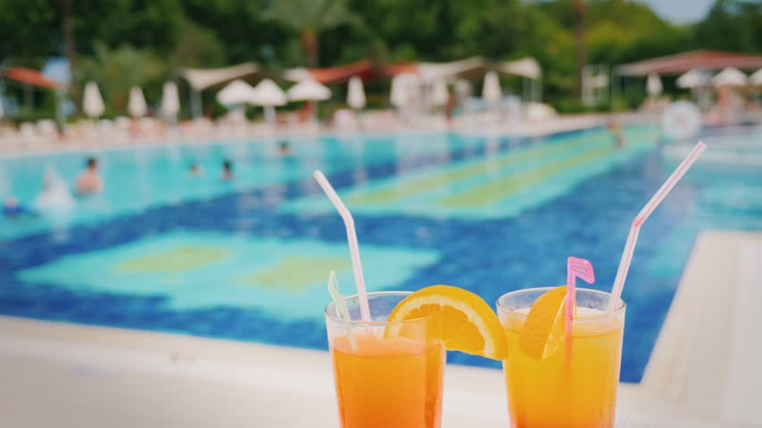 Refreshing alcoholic beverages served in glass tumblers with straws and orange slices placed on poolside deck. Bright blue swimming pool water in background provides vacation atmosphere.