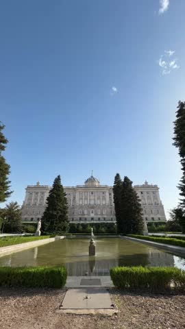 Majestic Royal Palace of Madrid seen from the tranquil Sabatini Gardens, featuring its neoclassical facade, a central fountain, and reflecting pool under a clear blue sky on a sunny day