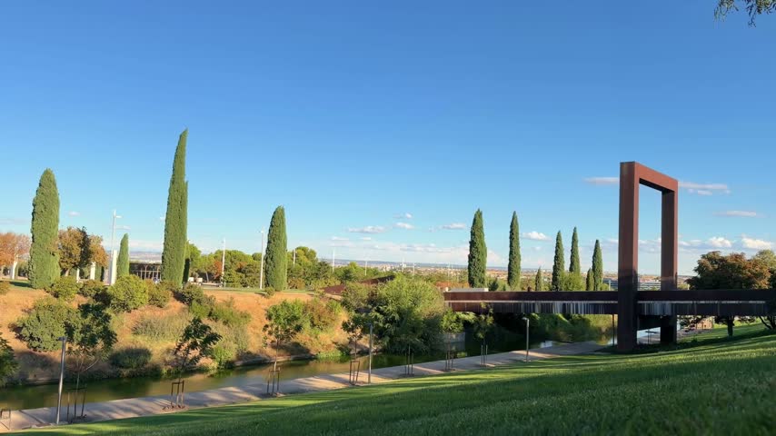 Beautiful landscape of the Parque Juan Carlos I in Madrid, with its distinctive rusted steel bridge over the creek, surrounded by lush green lawns, cypresses, and trees on a sunny day