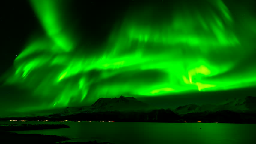 Time lapse of vibrant green aurora borealis swirling in night sky above snow capped mountains and water in Bentsjord, Troms, Norway
