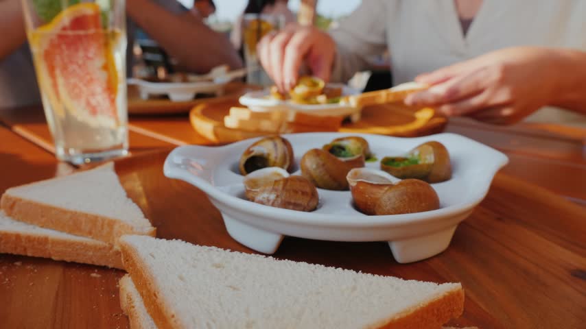 Close up shot focusing on white serving plate containing cooked escargot shells while person in background eats meal with toast in sunny outdoor dining environment.