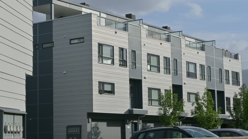 Static shot of a modern apartment building with balconies and trees in the foreground under a partly cloudy sky in Penticton, British Columbia.