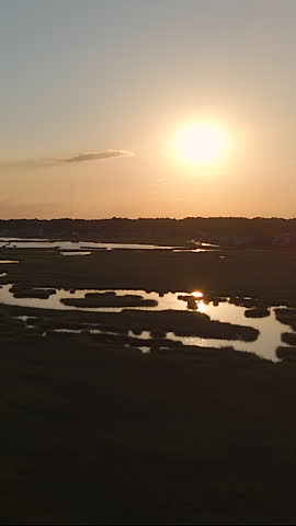 Vertical View Of Bright Sun Setting Over Coastal Salt Marsh Wetlands, Casting Warm Golden Reflections Across Calm Estuary Waters. - aerial shot. Assateague National Park.