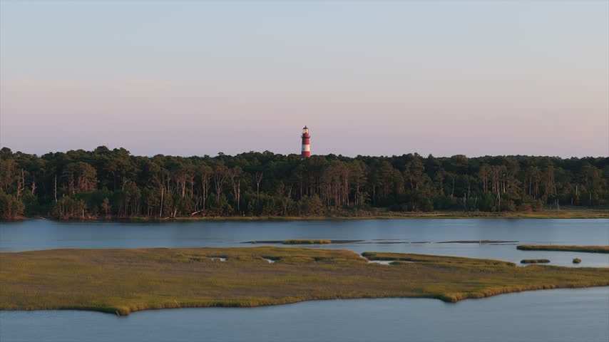 Lighthouse Rises Above Dense Forest Canopy Across Calm Salt Marsh Waterway At Soft Sunset Twilight. - aerial shot. Assateague, Maryland.