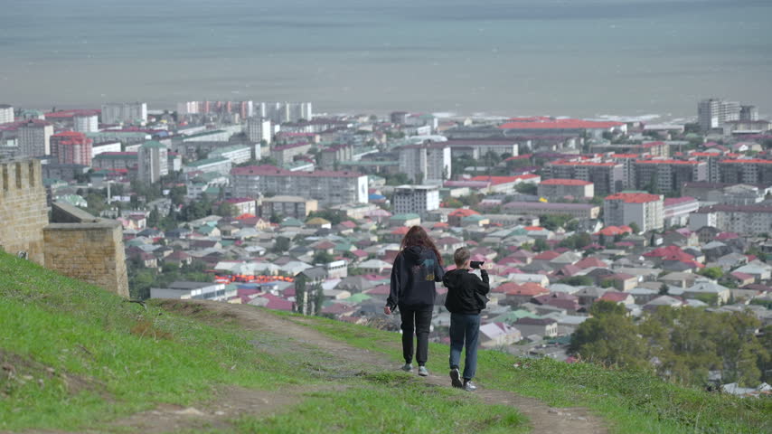 Two people walking downhill on grassy hillside overlook above dense coastal city, one holding phone toward skyline while viewing packed rooftops, distant sea, and hazy urban panorama