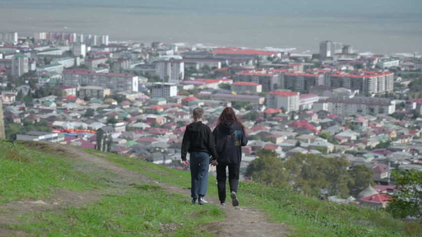 Two adults walking from behind along grassy hillside path above coastal cityscape, moving toward distant urban horizon under hazy daylight in calm panoramic travel mood