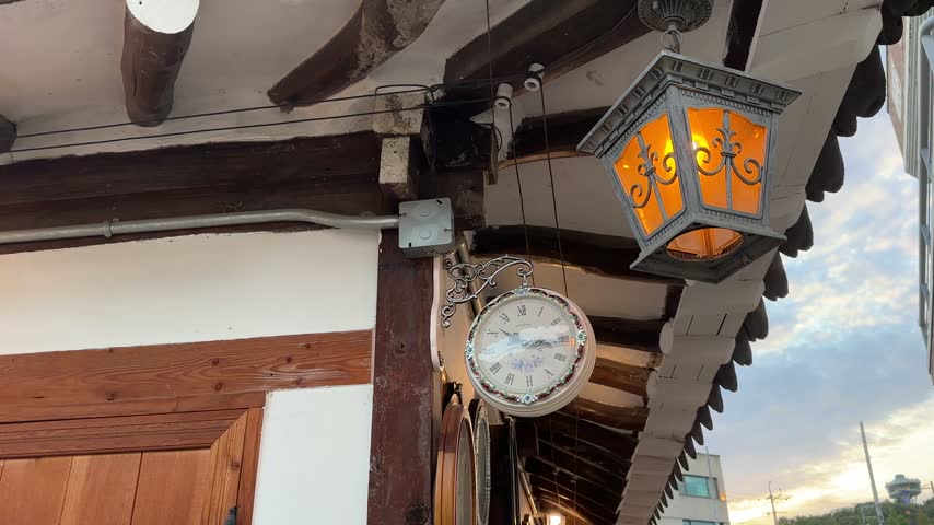 Glowing Orange Lantern Above Vintage Clock on Wooden Exterior
Exterior video footage focusing on a warm glowing lantern positioned above a vintage hanging clock beneath exposed wooden roof beams.