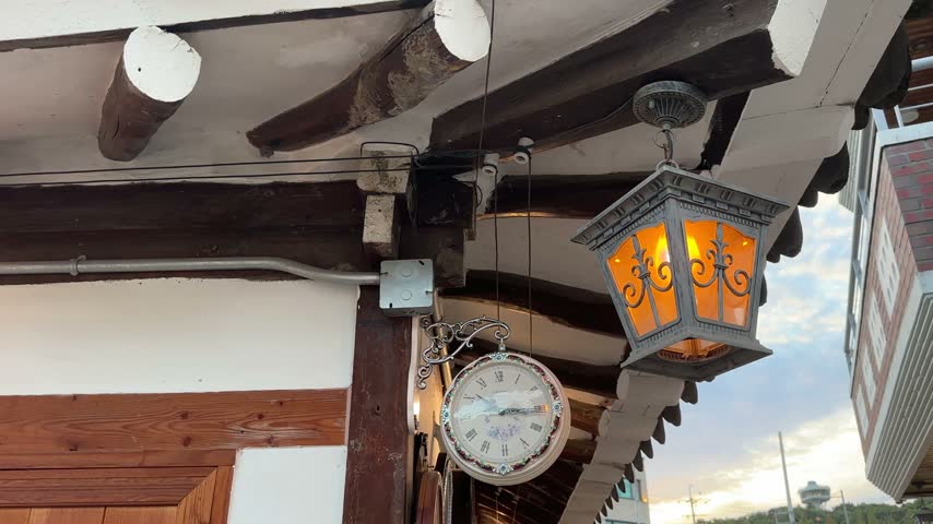 Close View of Vintage Clock and Glowing Hanging Lantern
Video footage featuring a vintage hanging clock beside a glowing orange lantern under exposed wooden roof beams, highlighting decorative exterior details.