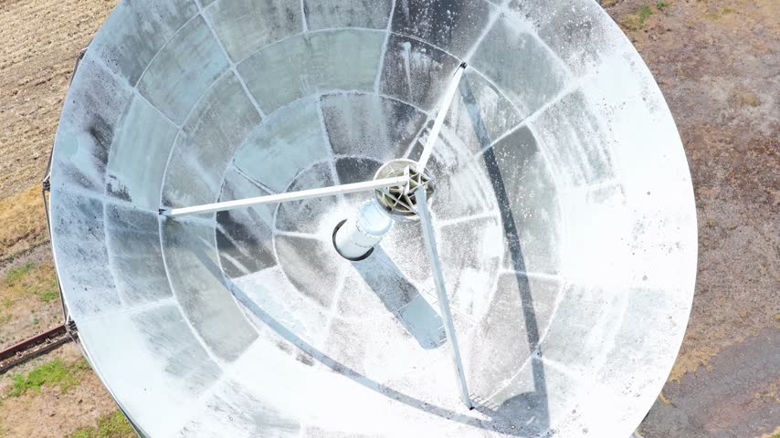 An overhead view of a large white satellite dish rotating in a rural field to track communication signals during the day.