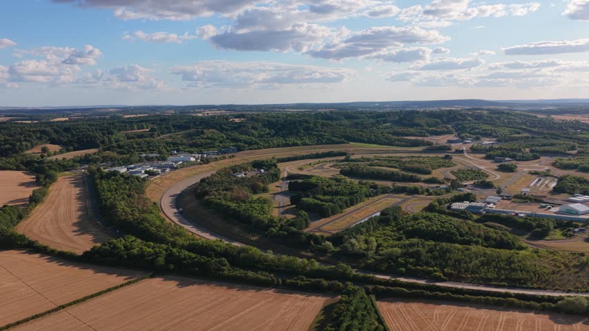 Wide drone perspective of a vehicle testing facility featuring complex road circuits surrounded by green forests and dry agricultural land under a clear blue sky during the late afternoon hours