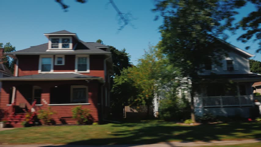 Panning shot from moving vehicle showing diverse residential architecture along quiet suburban street. Cinematic view of historic homes featuring classic porches and lush landscaping.