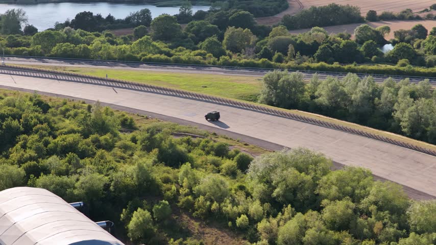 Black military vehicle maneuvers across a wide asphalt track surrounded by lush green trees and a lake during a sunny afternoon at a testing facility