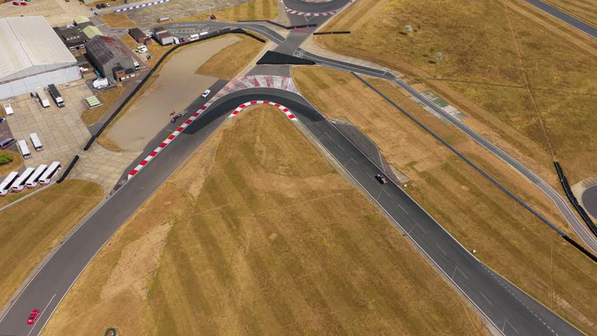 Aerial view of a race car navigating a tight bend at the Silverstone Circuit in England during a sunny day with dry grass visible around the asphalt track