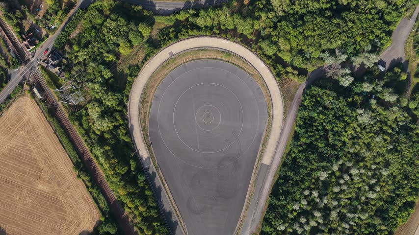 High altitude perspective looking down at a specialized automotive test track surrounded by dense green woodland and fields under bright daylight conditions