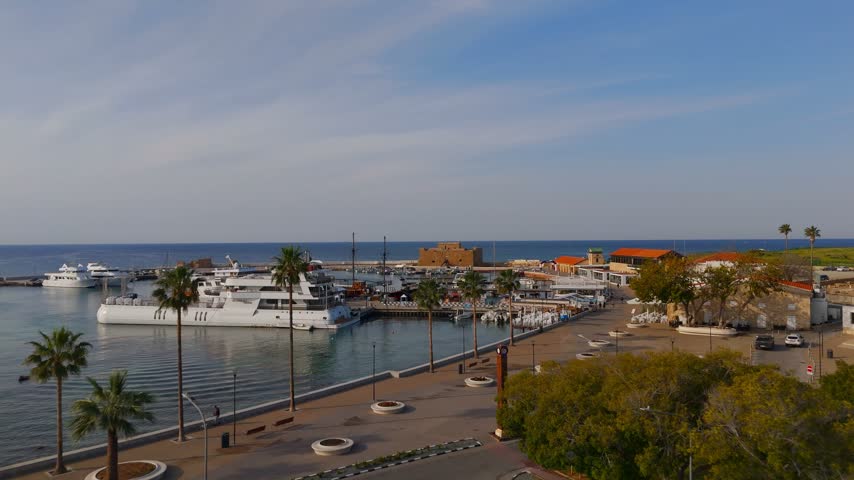 A low-altitude drone shot glides past palm trees and fishing boats towards the historic Paphos Medieval Castle at the harbor entrance in Cyprus.