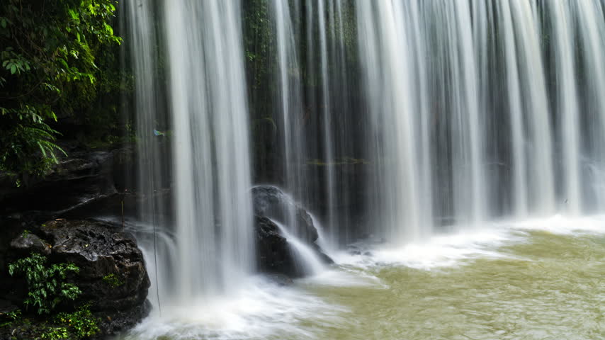 cinematic timelapse of a tropical waterfall flowing over dark moss-covered rocks surrounded by lush green rainforest vegetation. The flowing water creates a peaceful, refreshing natural atmosphere