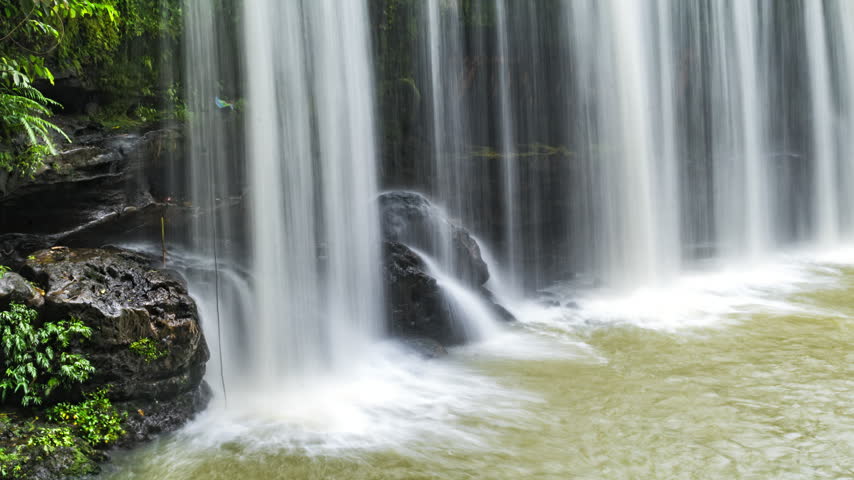 cinematic timelapse of a tropical waterfall flowing over dark moss-covered rocks surrounded by lush green rainforest vegetation. The flowing water creates a peaceful, refreshing natural atmosphere