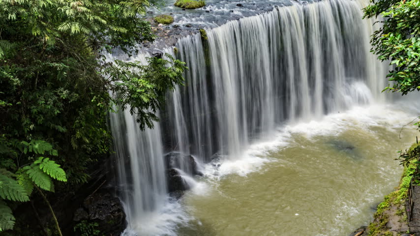 cinematic timelapse of a tropical waterfall flowing over dark moss-covered rocks surrounded by lush green rainforest vegetation. The flowing water creates a peaceful, refreshing natural atmosphere