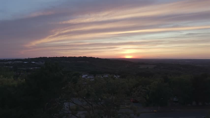 Aerial view of a golden sunset over wooded hills beneath streaks of pink clouds.