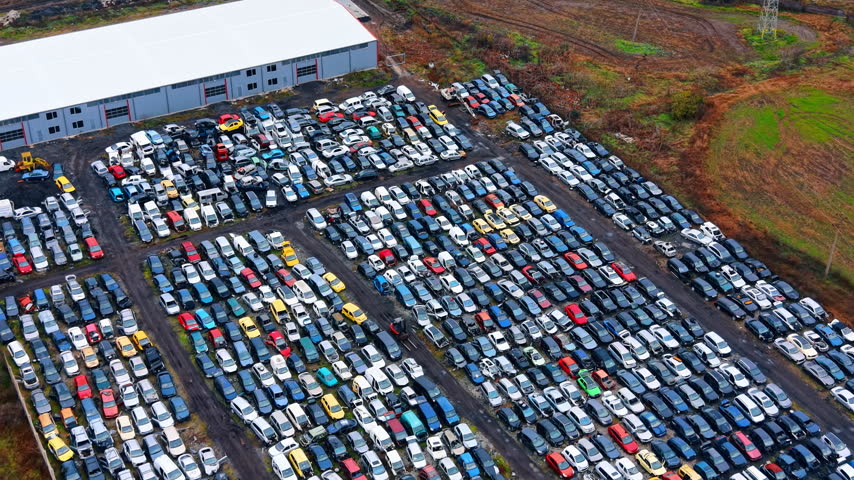 Diagonal aerial perspective of car scrap yard and warehouse. Rows of wrecked cars parked in a salvage facility near a large gray industrial building.