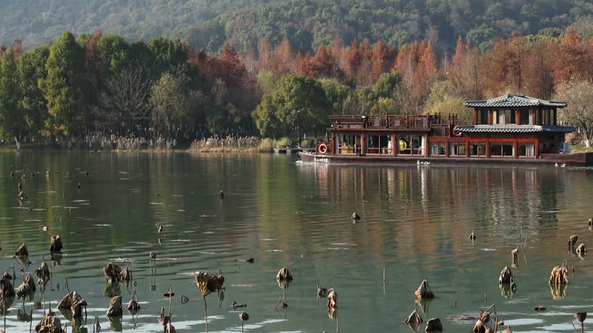A traditional Chinese tourist boat sailing on a calm lake surrounded by autumn foliage and dried lotus flowers.