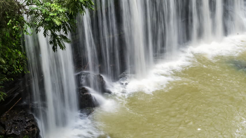 cinematic timelapse of a tropical waterfall flowing over dark moss-covered rocks surrounded by lush green rainforest vegetation. The flowing water creates a peaceful, refreshing natural atmosphere