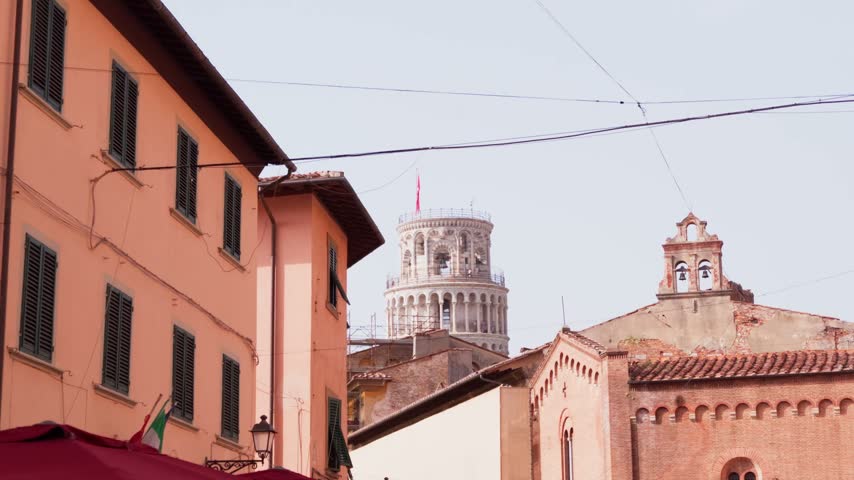 Slow gimbal walk past old town rooftops and brick church gable with Leaning Tower top peeking above in daylight, Pisa, Tuscany, Italy