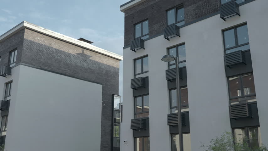 Low angle view of quiet urban apartment buildings with repeating windows, dark brick upper floors, light facades, and streetlamp under pale blue sky