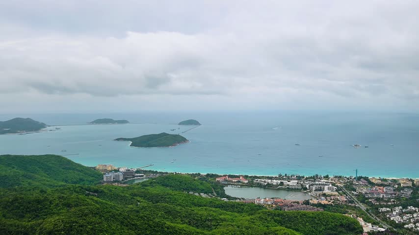 Yalong Bay panorama stretches across Hainan Island, with turquoise water, offshore islands and curving resort coastline. Elevated coastal scenery reveals green hills beneath cloudy skies.