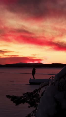 Sideways orbiting view past a snowy spruce branch toward a person on a jetty on a frozen lake at red sunset, Finnish Lapland, Finland