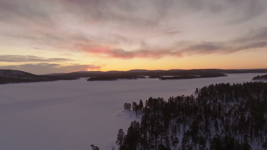 Slow descending view over a forested shore and frozen lake with pink polar twilight glow on the horizon, Finnish Lapland, Finland