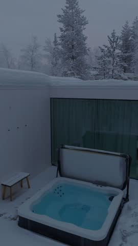 Low static view of an open hot tub beside a glass-walled cabin in snowy forest at polar twilight, Finnish Lapland, Finland