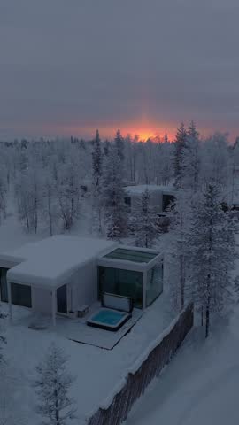 Slow descending approach toward a glass cabin with hot tub and sunrise glow behind frosted trees, Finnish Lapland, Finland