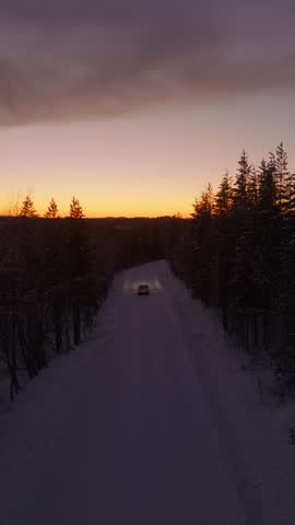 Low forward flight over a snowy forest road toward a car with headlights at orange dusk, Finnish Lapland, Finland