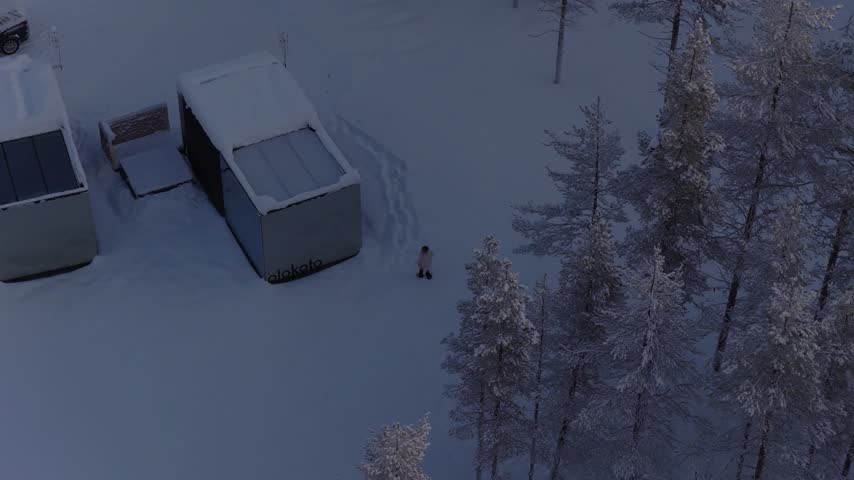 Top-down rotating view of two snow-covered mirror cabins with a person standing in deep snow at dusk, Finnish Lapland, Finland