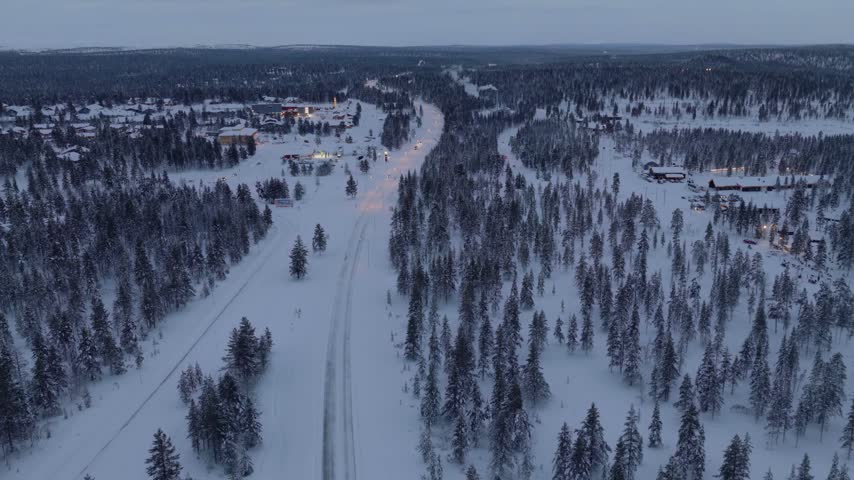 Flying forward over snowy taiga forest along a lit road toward an illuminated village at dusk, Finnish Lapland, Finland