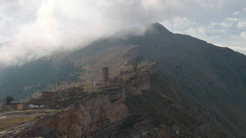 Cloud-wrapped mountain ridge with weathered stone ruins along cliff edge and drifting mist over steep rocky slopes in soft daylight