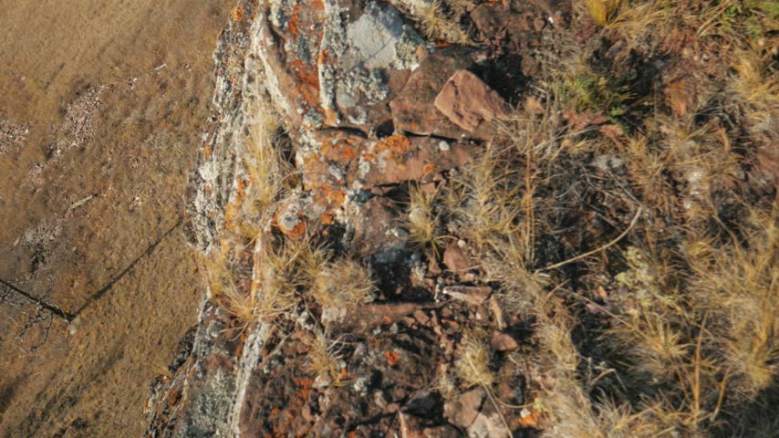 A person in yellow boots walks along the edge of a steep mountain made of red Devonian sandstone. A first-person view of the autumn landscape features dry yellow grass and a vast field below in Khakassia, Siberia.
