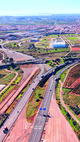Motorway junction toward Valencia and Alicante, Spain. Road markings, blue signs, cars and trucks on multilane lanes, a bridge crossing, orchards and a coastal town. Vertical video.